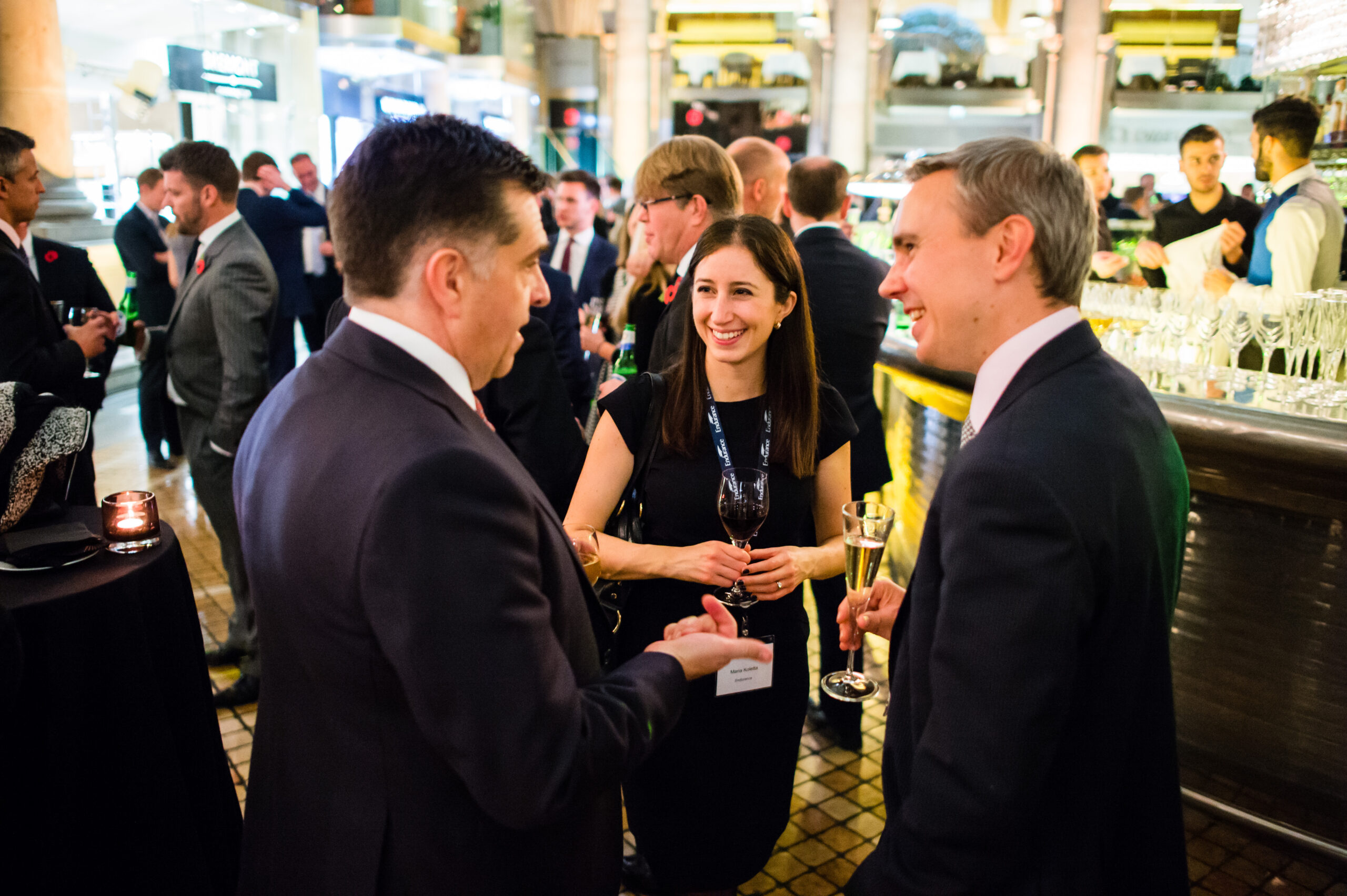 A group of professionally dressed people interact at a social event in a well-lit venue. A woman in a black dress holds a wine glass, smiling, while two men in suits engage in conversation. Other attendees are visible in the background near the bar. James Gifford-Mead Photography - Event Photographer London