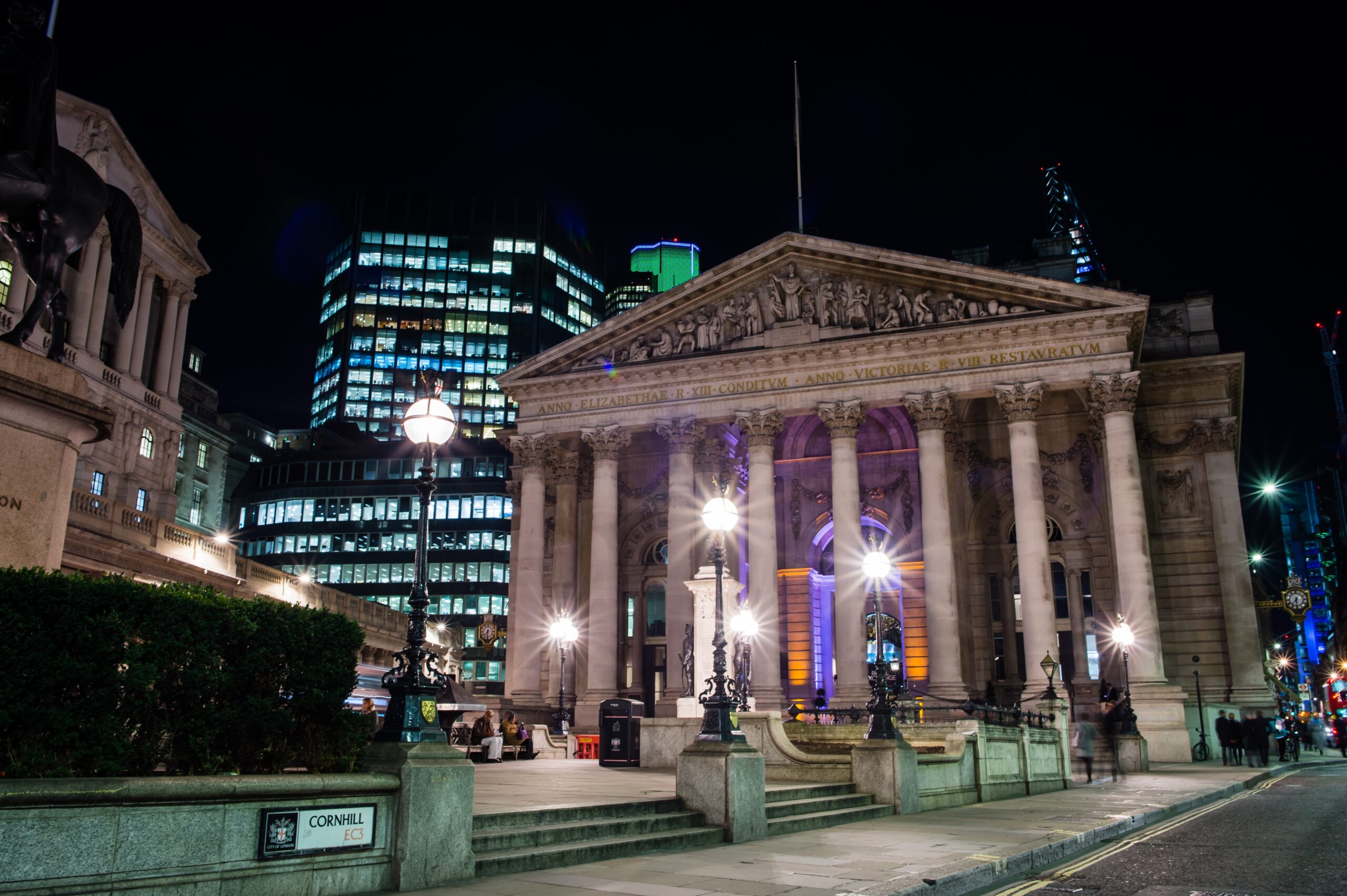 A grand, illuminated classical building stands at night, surrounded by modern skyscrapers with glowing windows. The scene features street lamps and steps leading to the entrance, creating a contrast between historic and contemporary architecture. James Gifford-Mead Photography - Event Photographer London