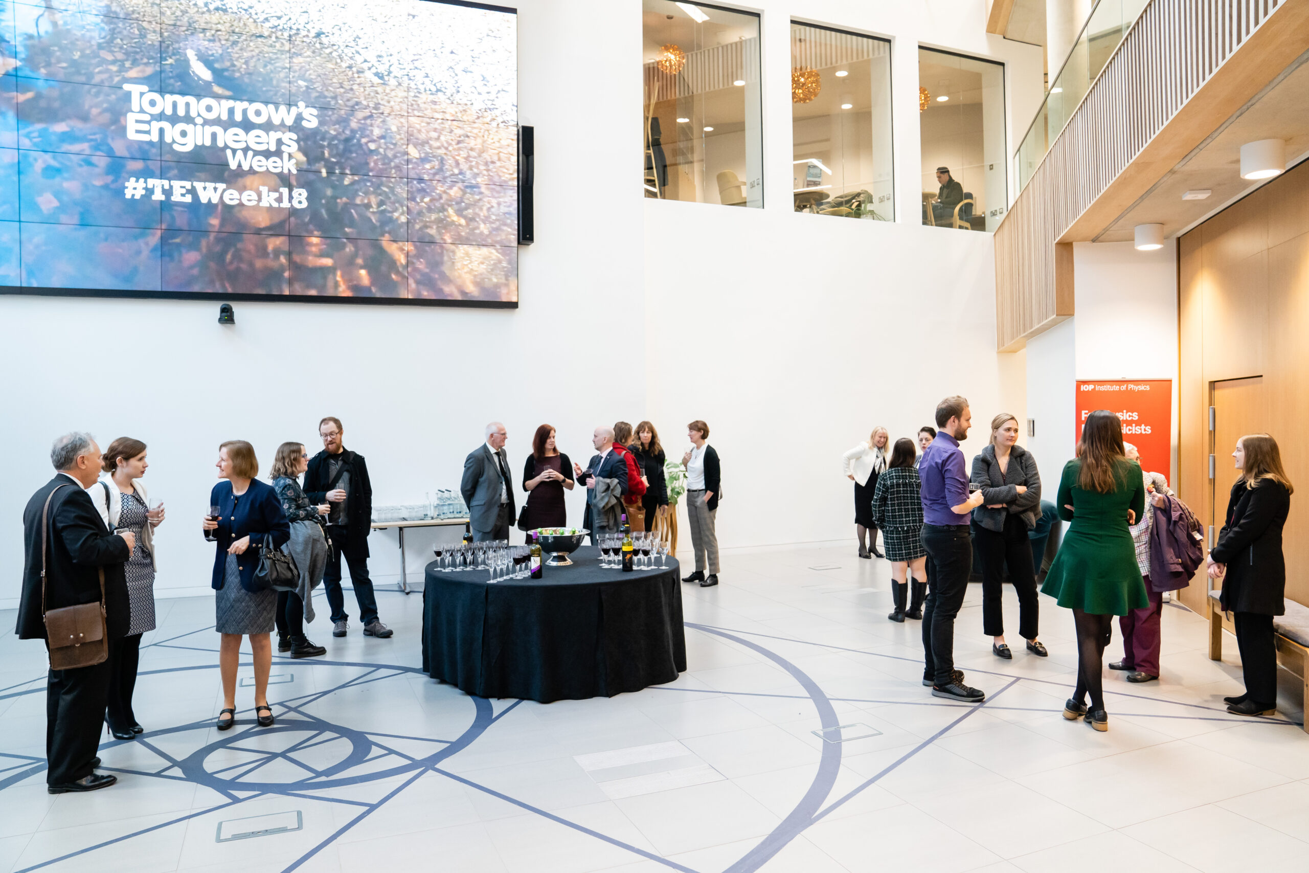 A group of people in a modern building lobby attend an event. A screen displays "Tomorrow's Engineers Week #TEWeek18." Attendees are mingling around a circular refreshment table set with drinks and glasses. James Gifford-Mead Photography - Event Photographer London
