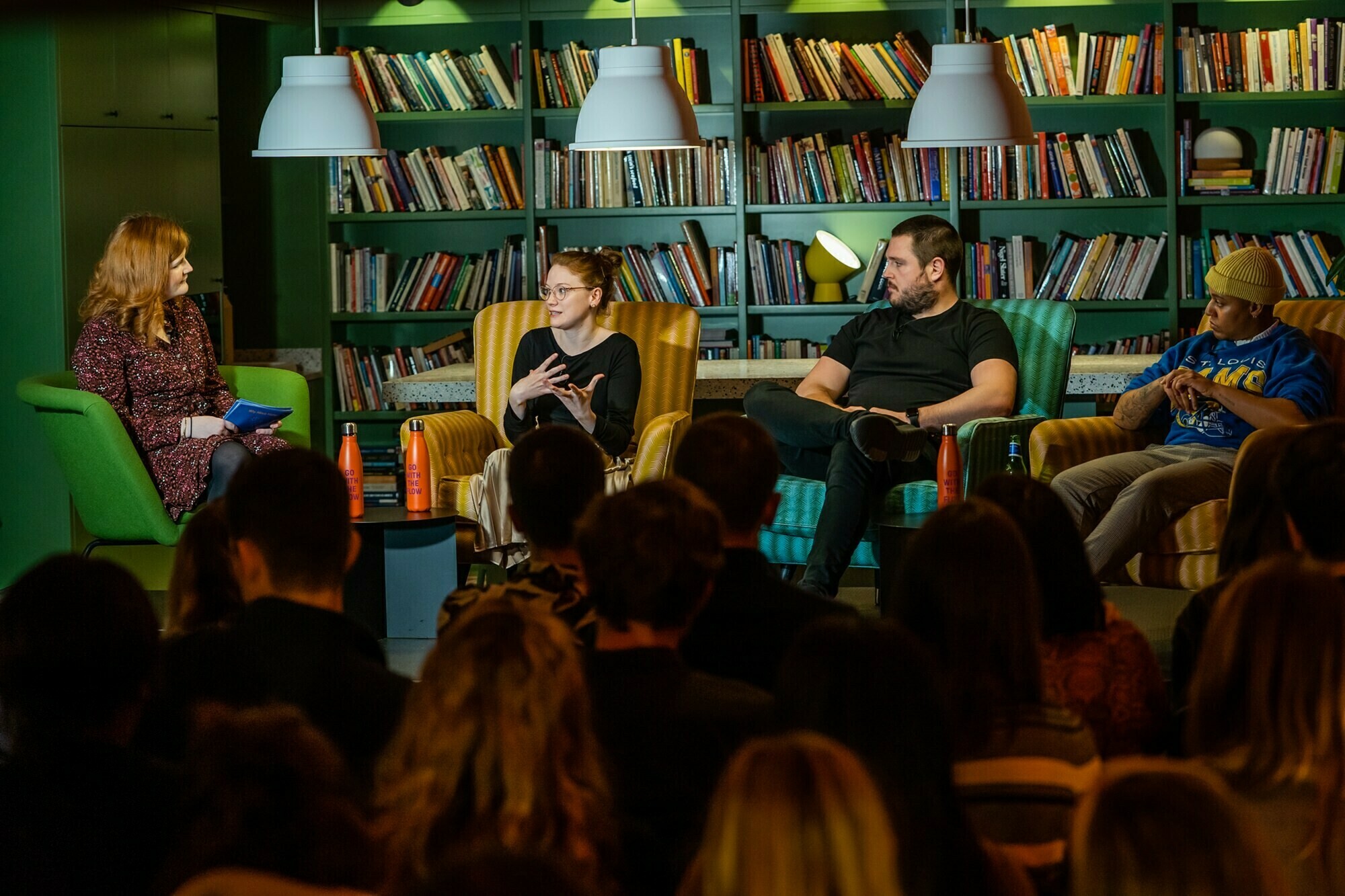 A panel of three people sits in armchairs on a stage, engaged in discussion. A fourth person is moderating, seated to the left. An audience listens attentively. Bookshelves and pendant lights are visible in the background. James Gifford-Mead Photography - Event Photographer London