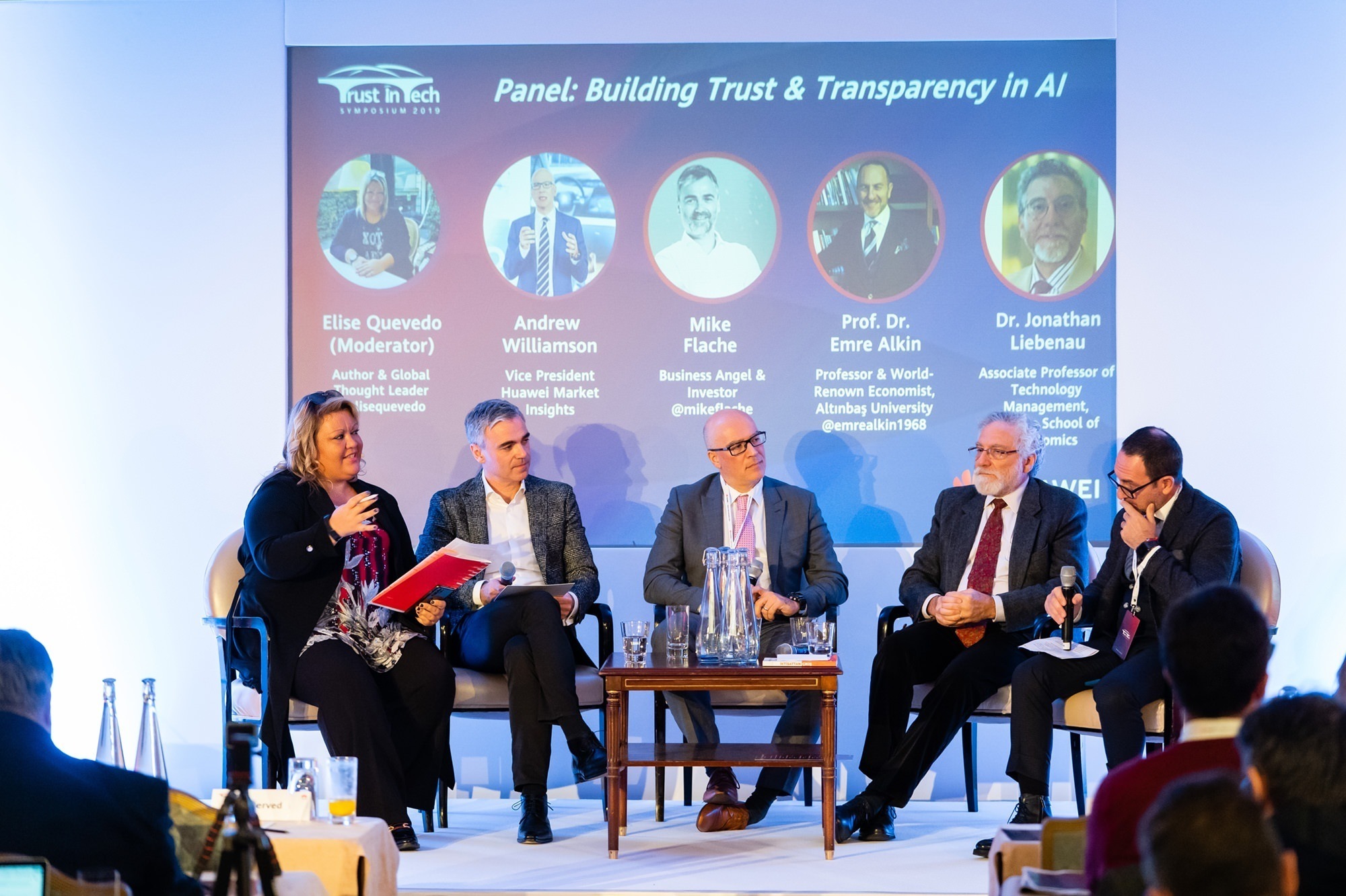 Five individuals are seated on a stage having a panel discussion titled "Building Trust & Transparency in AI." There is a screen in the background displaying their names and titles. The moderator is holding a red folder. James Gifford-Mead Photography - Event Photographer London