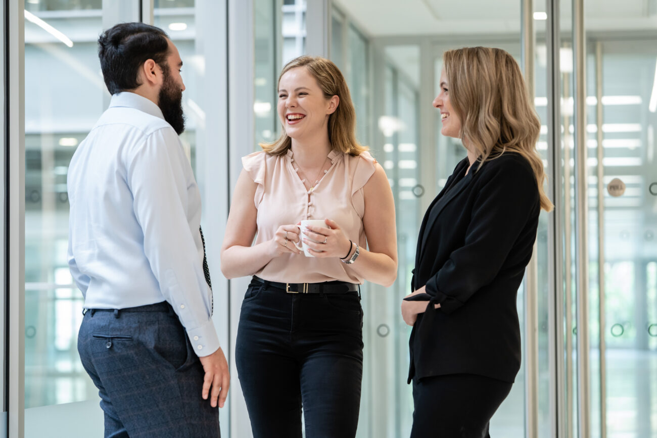 Three colleagues stand in a bright office hallway, engaged and smiling, as captured by a corporate photographer in London. One man and two women, dressed in business attire, suggest a professional environment. Glass walls and doors frame their pleasant conversation