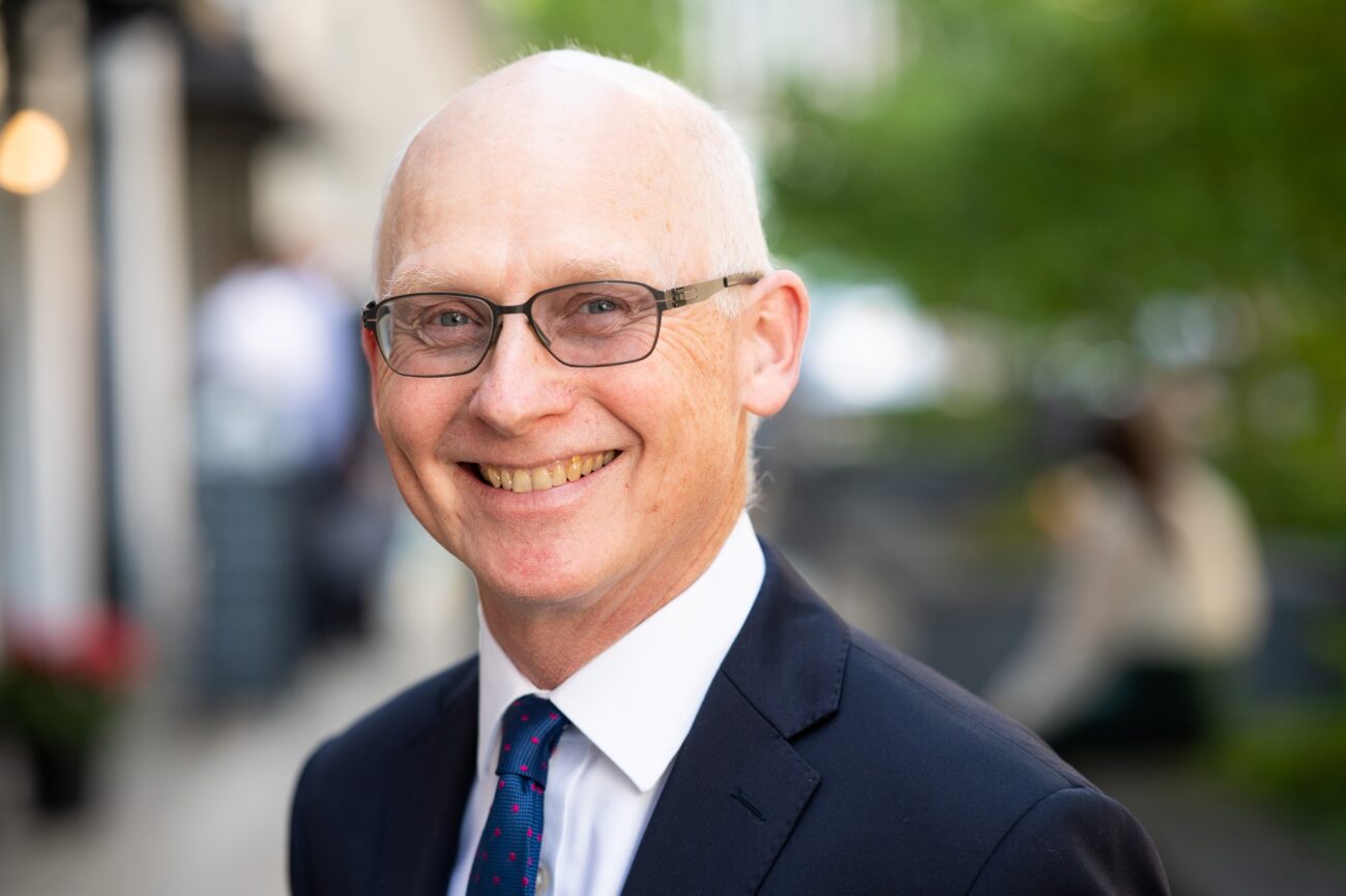 A bald man wearing glasses and a dark suit with a dotted tie smiles broadly. The background is a blurred outdoor setting with greenery and a person sitting on a bench. James Gifford-Mead Photography - Event Photographer London