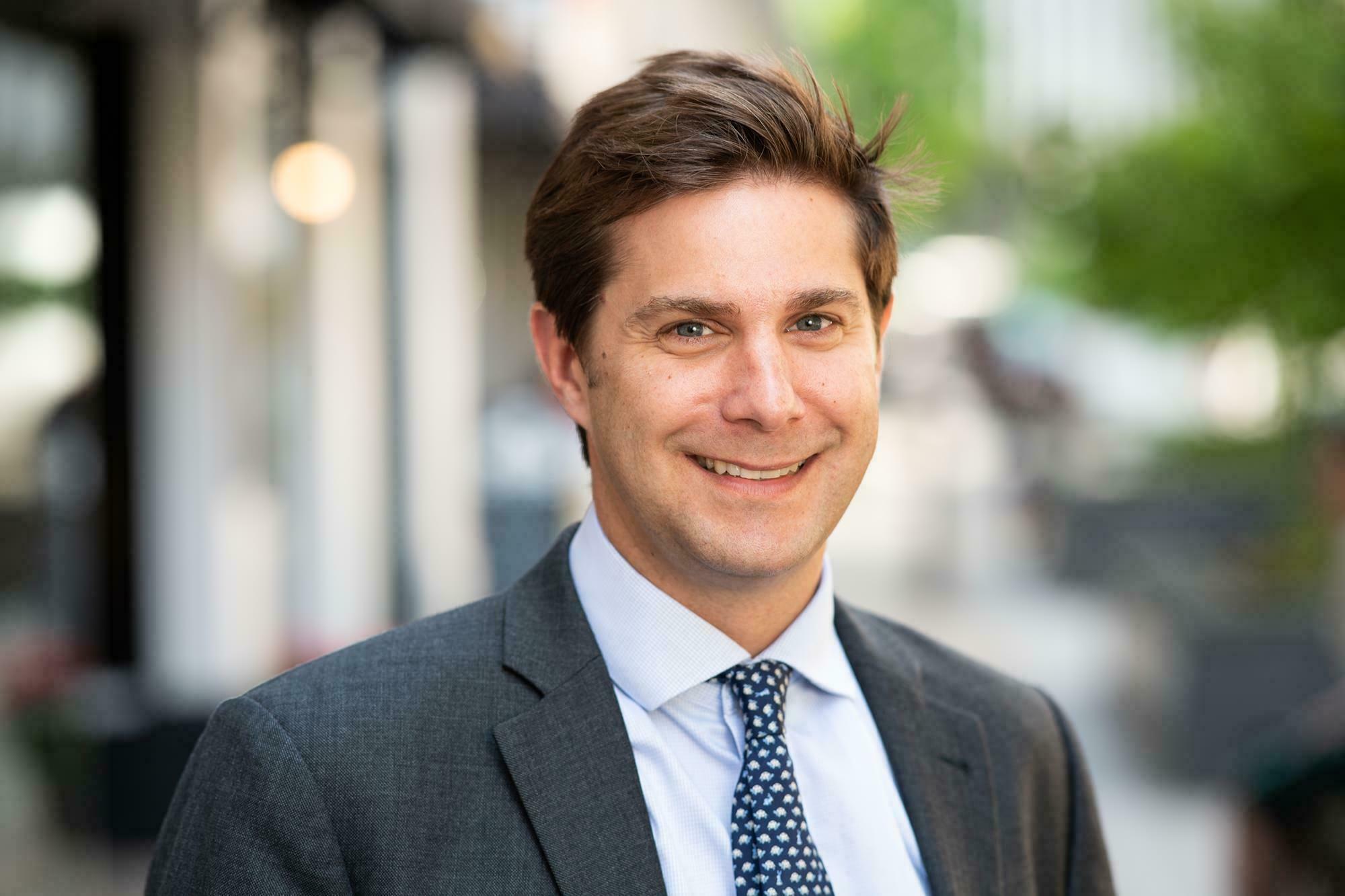 Smiling man in a suit and tie stands outdoors with a blurred street and greenery in the background. James Gifford-Mead Photography - Event Photographer London