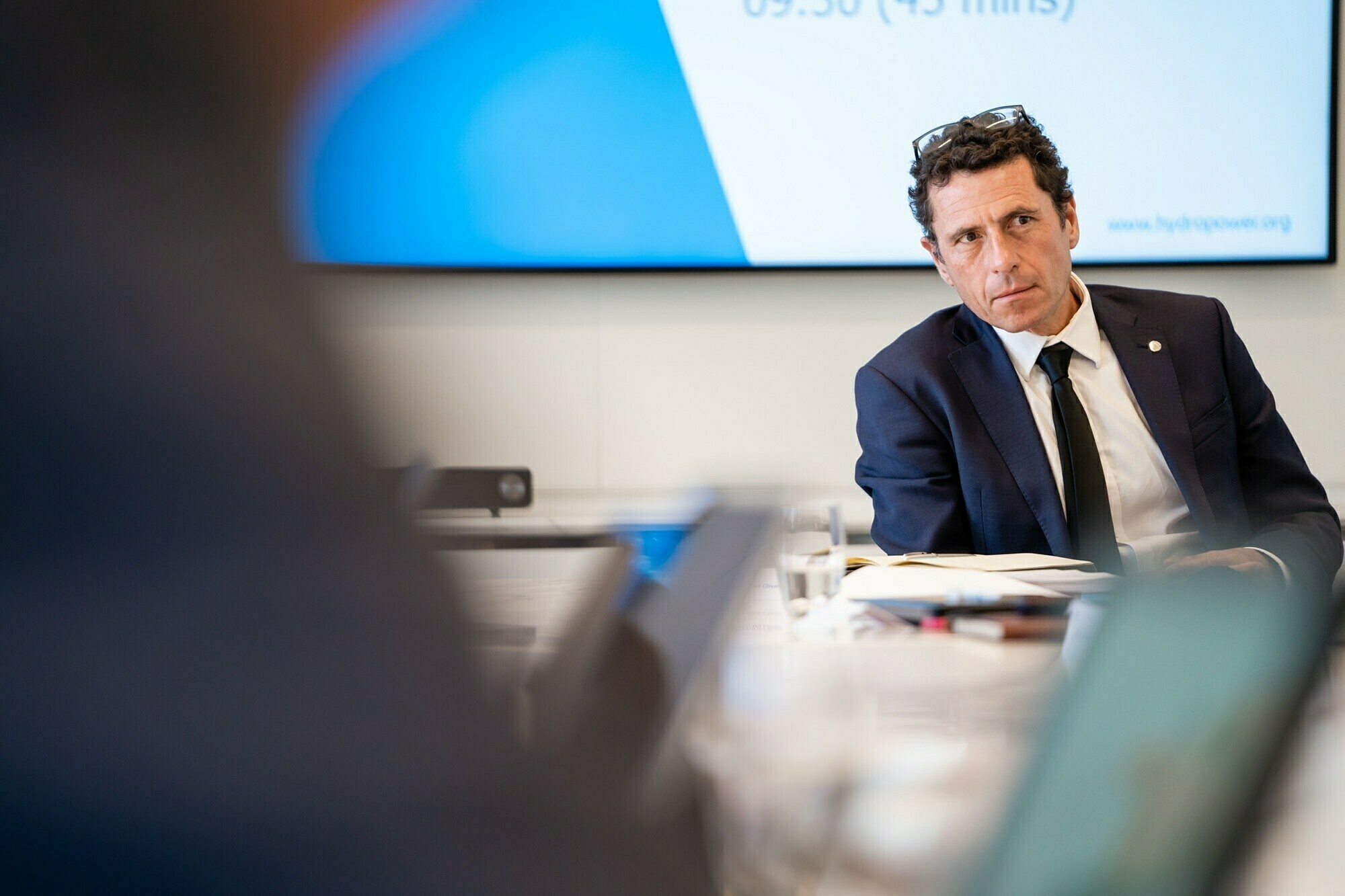 A man in a dark suit sits at a conference table, appearing engaged in a discussion. He has glasses on his head and a notepad in front of him. A presentation screen is visible in the background. The focus is on him as he listens attentively. James Gifford-Mead Photography - Event Photographer London
