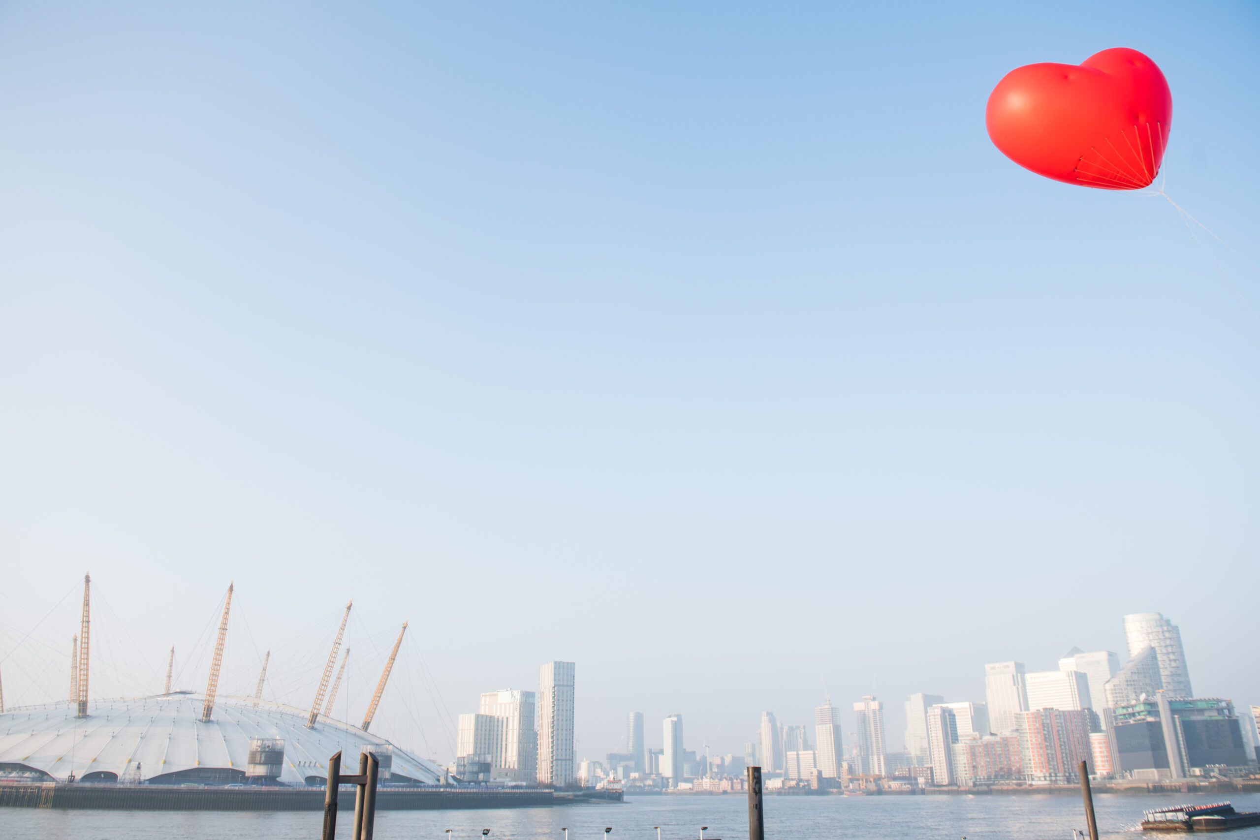 A vibrant red heart-shaped balloon floats in a clear blue sky above a cityscape, captured brilliantly by a corporate photographer in London. Below, modern skyscrapers and a dome structure adorn the waterfront, creating a lively urban backdrop. James Gifford-Mead Photography - Event Photographer London
