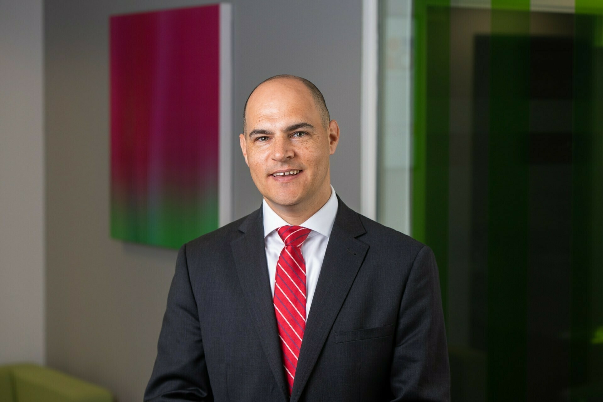 A man in a suit and red tie stands indoors, smiling. The background features a modern design with a colorful gradient art piece on the wall. The setting appears professional and well-lit. James Gifford-Mead Photography - Event Photographer London