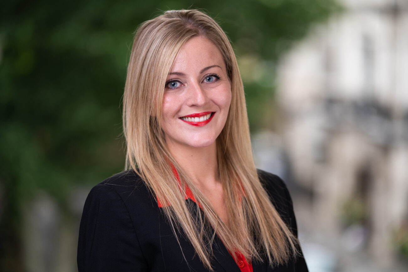 A person with long blonde hair and a black blazer stands outdoors, capturing the corporate headshot photographer's perfect moment. They smile brightly with red lipstick, set against a backdrop of greenery and blurred buildings. James Gifford-Mead Photography - Event Photographer London