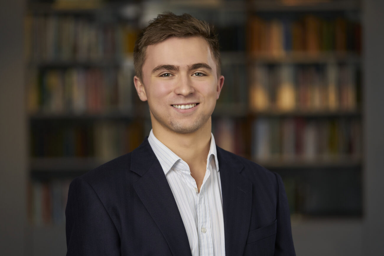 A person in a dark suit and light striped shirt smiles at the camera, captured perfectly by a corporate headshot photographer, with a blurred background of bookshelves adding depth to the image. James Gifford-Mead Photography - Event Photographer London