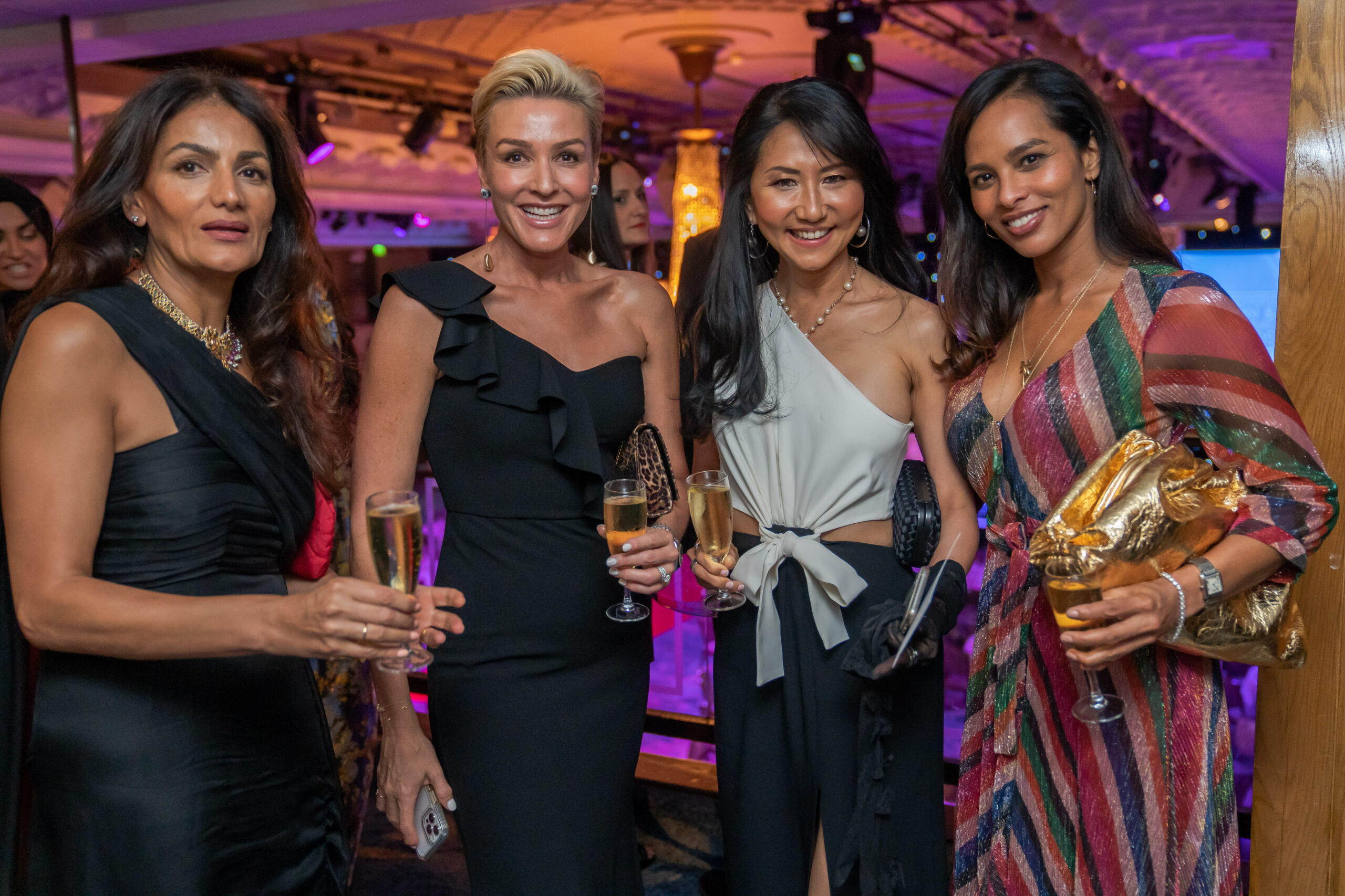 Four women in elegant dresses stand together, smiling, and holding glasses of champagne at an upscale indoor event. The venue has warm lighting and a decorated ceiling, suggesting a celebratory atmosphere. James Gifford-Mead Photography - Event Photographer London