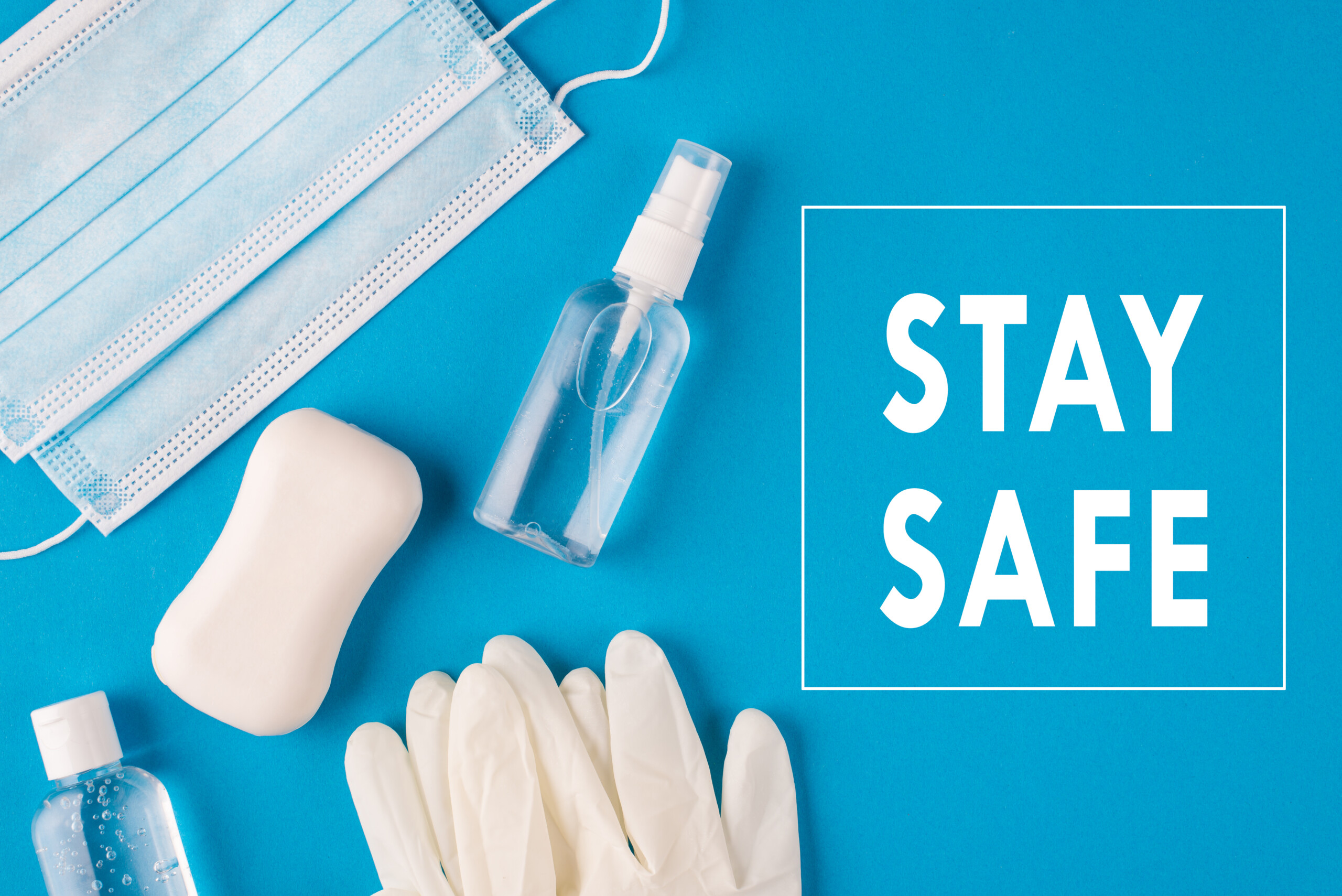 A flat lay of a blue background with items for hygiene and safety: a face mask, a bar of soap, a small sanitizer bottle, and a pair of gloves. The words "STAY SAFE" are printed in white on the right side. James Gifford-Mead Photography - Event Photographer London