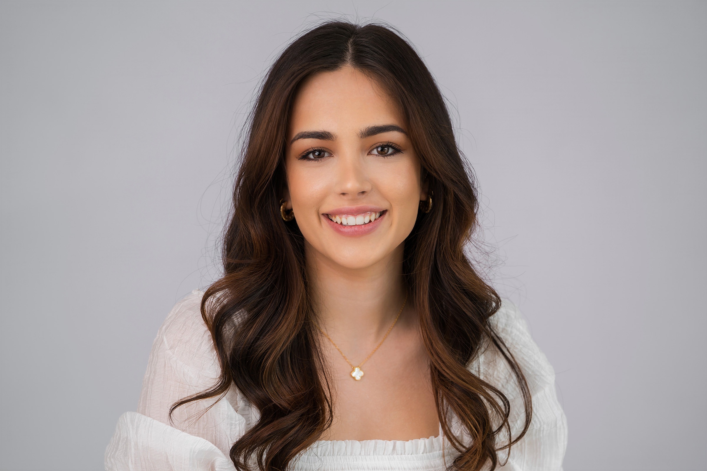 Smiling woman with long, wavy dark hair. She wears a white blouse and a gold necklace with a small pendant. The background is a light gray, providing a clean and simple backdrop. James Gifford-Mead Photography - Event Photographer London
