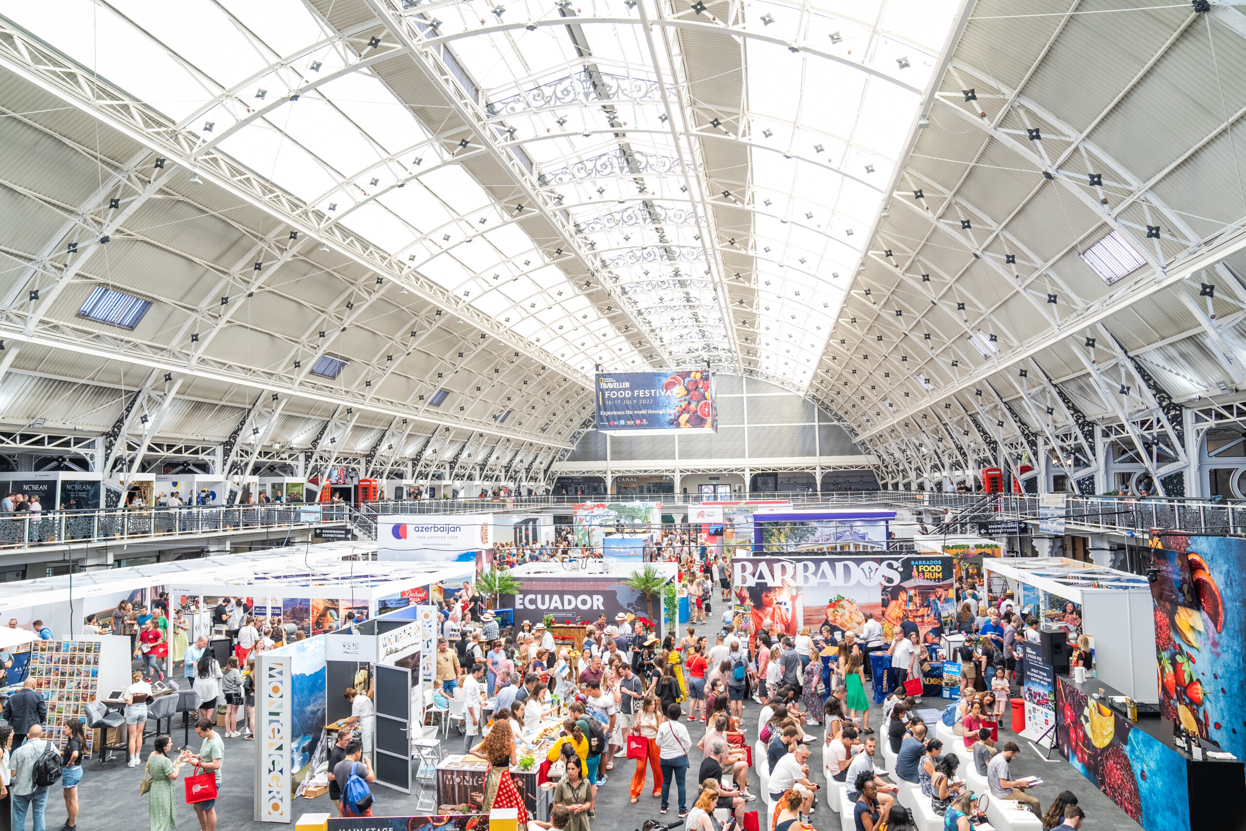 A large, bustling food festival inside a spacious exhibition hall. Visitors explore various international food stalls with banners from different countries, including Ecuador and Barbados. The hall's arched ceiling has large skylights, filling the space with light