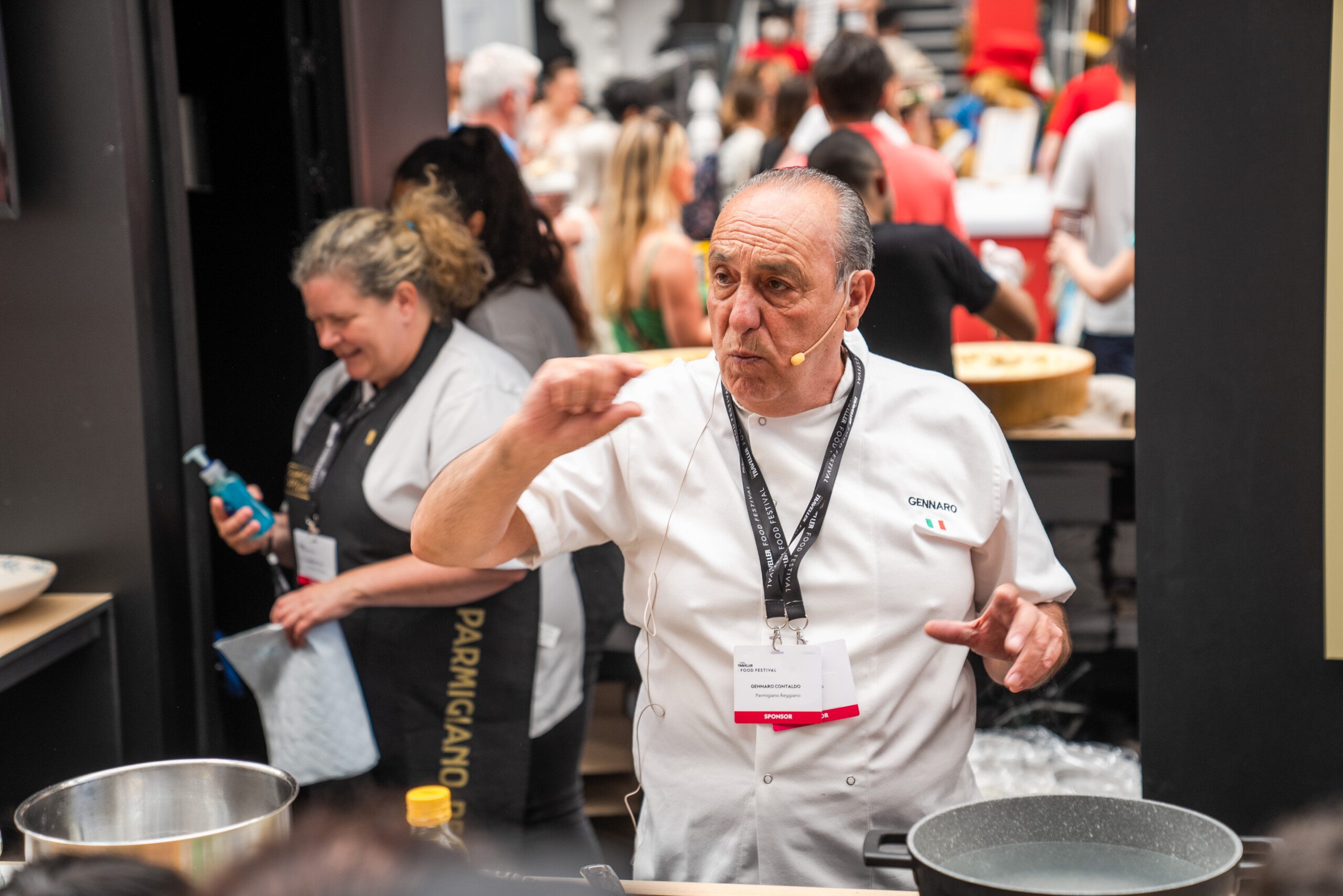 A chef wearing a white uniform and microphone gestures while speaking at a cooking event. Behind him, a woman in a black apron and several attendees are visible. Cooking equipment and ingredients are on the counter in front of the chef. James Gifford-Mead Photography - Event Photographer London
