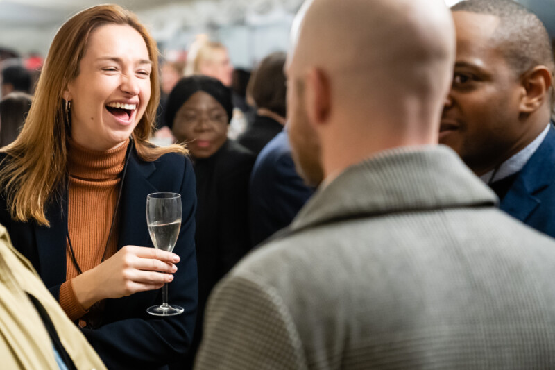 At a bustling London conference, a woman with long hair and a brown turtleneck laughs while holding a champagne glass. The photographer captures the lively atmosphere as several attendees in formal attire engage in animated conversation around her. James Gifford-Mead Photography - Event Photographer London