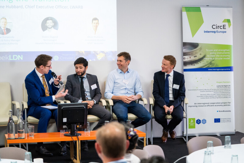 Four men in business attire are seated in a panel discussion at the London Conference. One speaks into a microphone while others listen attentively. Behind them is a screen and a CircE Interreg Europe roll-up banner, captured by a professional photographer. A table with glasses and a bottle is in the foreground. James Gifford-Mead Photography - Event Photographer London
