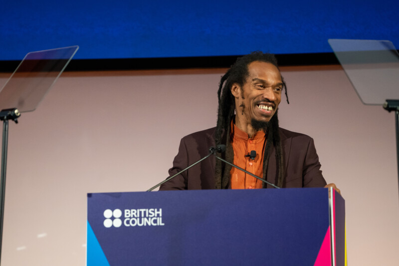 A man with long dreadlocks smiles at the podium, marked by the British Council logo. Captured by a London conference photographer, he's dressed in a dark blazer over an orange shirt, set against a blue and white backdrop with two transparent teleprompters on each side. James Gifford-Mead Photography - Event Photographer London