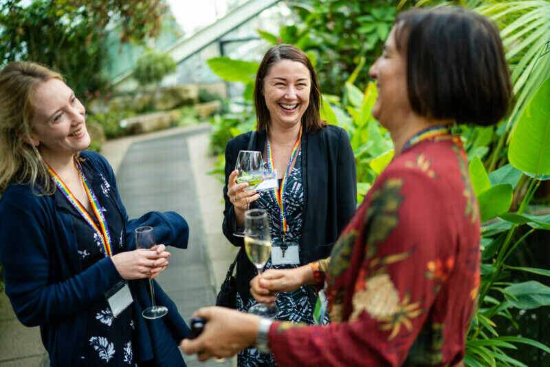 Three people smiling and holding drinks, engaged in conversation. They stand in a lush indoor garden with vibrant green plants, perfectly captured by a London conference photographer. Two are wearing lanyards, while one is holding a wine glass. James Gifford-Mead Photography - Event Photographer London