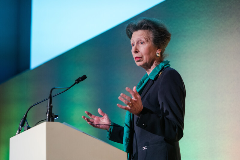 An older woman with short gray hair and a green scarf speaks at a podium, microphones poised to capture every word. The backdrop is a gradient of green and blue, reminiscent of the London Conference. She gestures animatedly, embodying the finesse of an experienced photographer. James Gifford-Mead Photography - Event Photographer London