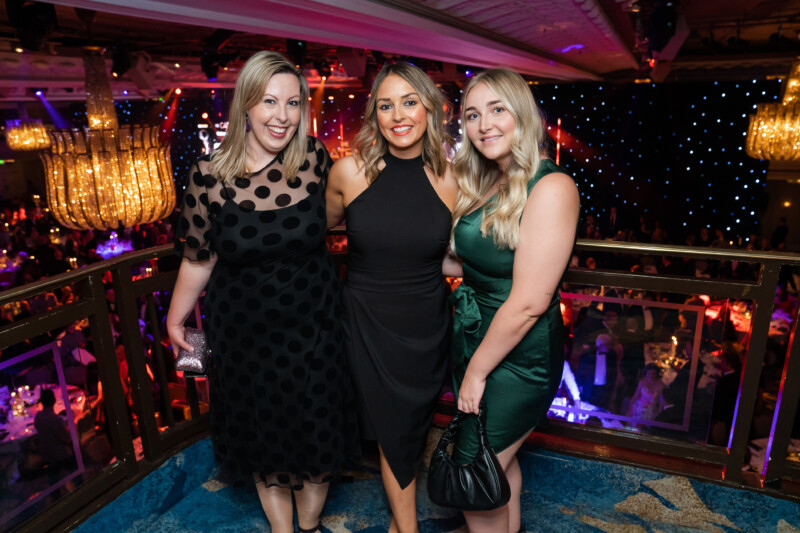 Three women in formal attire stand on a balcony at a London conference, capturing the essence of the event's elegance. The venue glistens with large chandeliers and tables set below with red lighting. They smile at the camera, with the central woman wearing a stunning black dress. James Gifford-Mead Photography - Event Photographer London