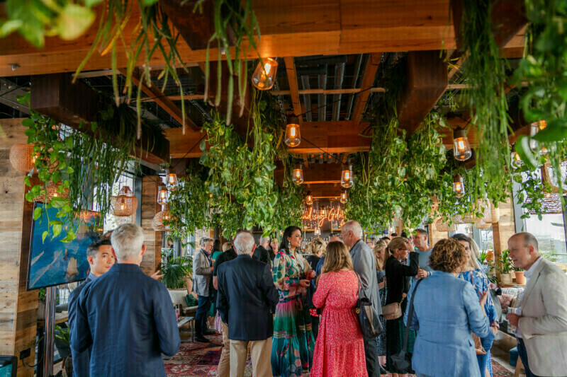 A lively indoor gathering captured by a London conference photographer. The space features a wooden ceiling adorned with hanging green plants and pendant lights, while attendees in diverse outfits engage in conversation amid the warm, inviting atmosphere. James Gifford-Mead Photography - Event Photographer London