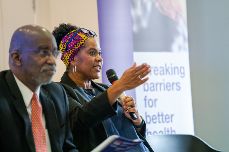 Two people sit side by side on a stage at the London Conference. One, wearing a suit and tie, sits quietly. The other, in a black jacket and colorful headwrap, speaks into a microphone against a backdrop sign reading "Breaking barriers for better health," as the photographer captures the moment. James Gifford-Mead Photography - Event Photographer London