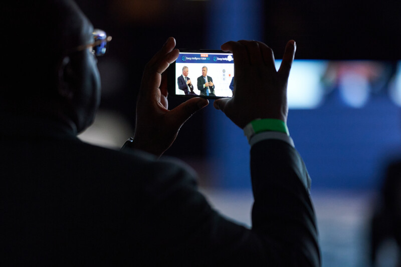 A London Conference photographer captures a photo of two individuals speaking on stage. The background is dark, and the screen displays the speakers clearly, their gestures visible. The photographer holding the smartphone is silhouetted against the scene. James Gifford-Mead Photography - Event Photographer London