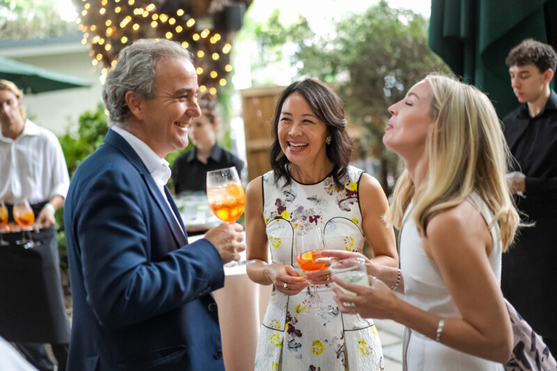 Three people in formal attire laugh and talk while holding drinks at an outdoor gathering. Captured by a skilled London conference photographer, string lights and trees create a festive atmosphere, with others in the background contributing to the lively setting. James Gifford-Mead Photography - Event Photographer London