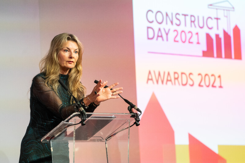 A woman with long blonde hair stands at a clear podium, speaking at a formal event, the "Construct Day 2021 Awards," captured by a skilled London Conference Photographer. Her dark outfit complements the professional ambiance. James Gifford-Mead Photography - Event Photographer London