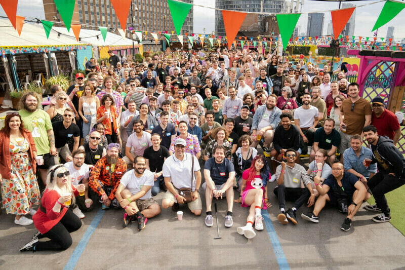 A large, diverse group of people gathers for a rooftop event in the heart of London. They are smiling at the camera, surrounded by colorful bunting and tall buildings in the background. The atmosphere is festive and lively, perfectly captured by a skilled conference photographer. James Gifford-Mead Photography - Event Photographer London