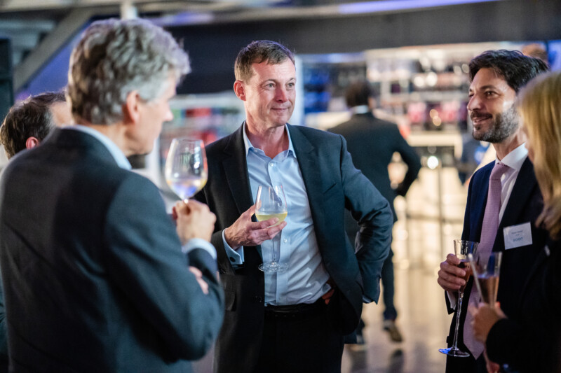 At a London conference, a group in formal attire mingles while a man at the center holds a wine glass. The photographer captures guests around him, all engaged in animated conversation, their drinks held aloft amidst the softly blurred backdrop of this elegant social gathering. James Gifford-Mead Photography - Event Photographer London