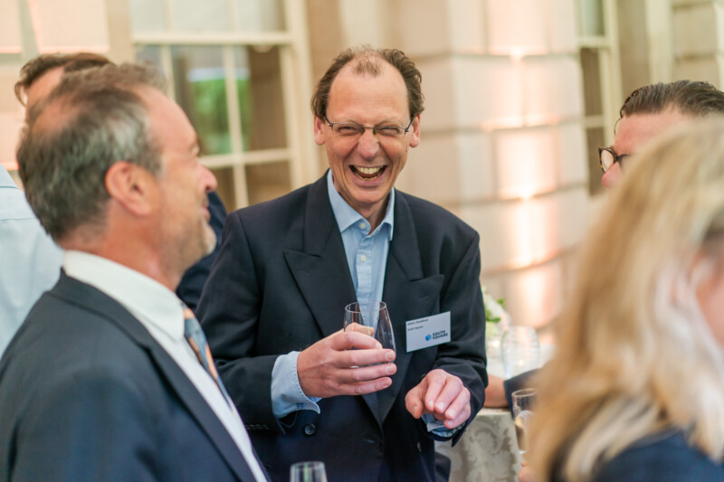 A man in a suit and glasses smiles while holding a glass, chatting with a group at an indoor event captured by a London conference photographer. The softly lit background reveals large windows, and other attendees are partially visible. James Gifford-Mead Photography - Event Photographer London