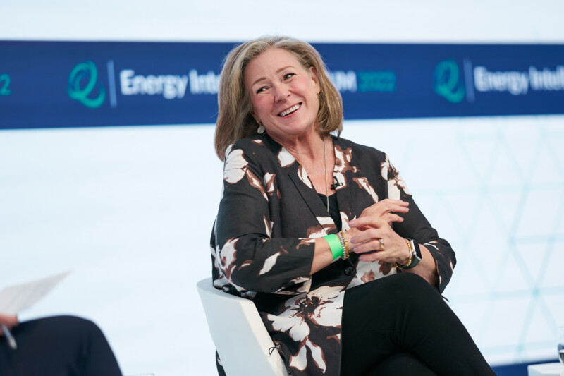 A smiling woman with light brown hair, dressed in a black floral top, sits on a stage during the Energy Intelligence Forum 2023. Captured by a London conference photographer, a banner displaying the forum's name is visible in the background. James Gifford-Mead Photography - Event Photographer London