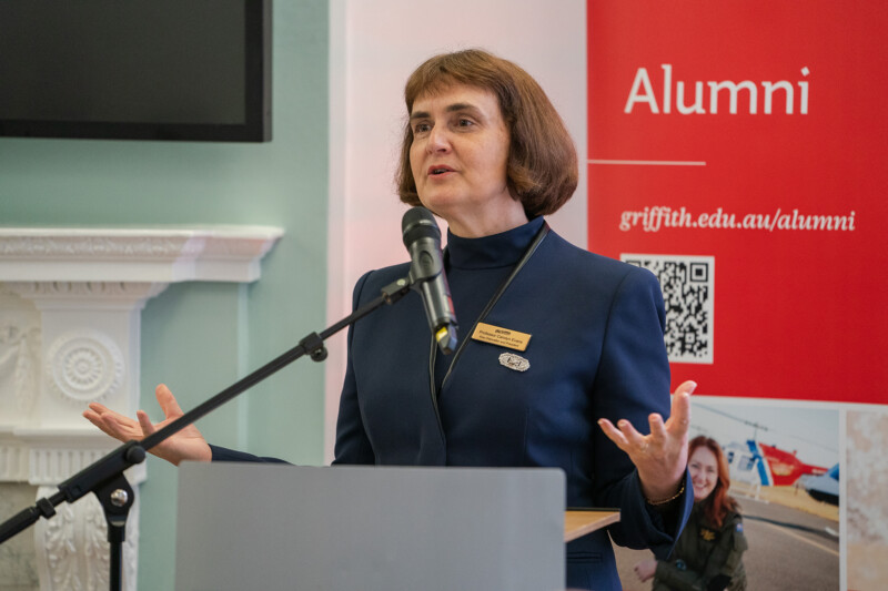 A woman in a navy suit speaks at a podium with a microphone. Behind her, a red banner reads "Alumni" alongside a website link. Captured by a skilled London Conference Photographer, she appears engaged, gesturing with her hands during the speech. James Gifford-Mead Photography - Event Photographer London