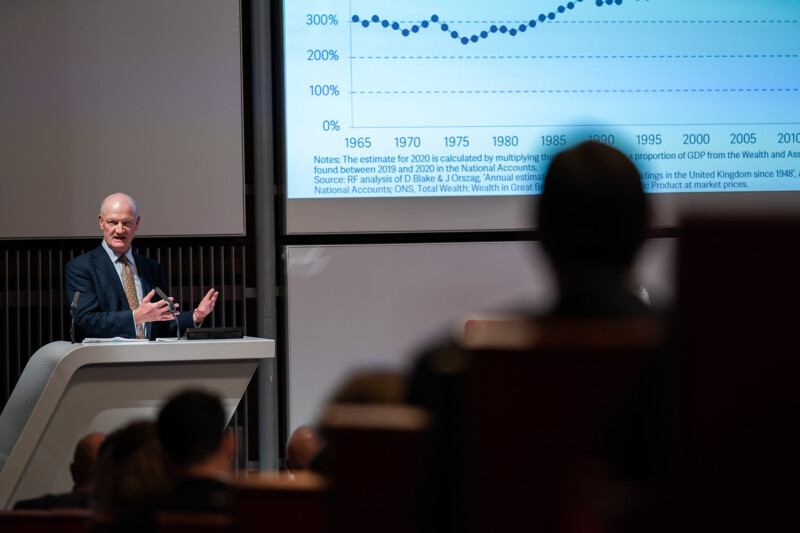 A speaker at a podium addresses an audience in a lecture hall, captured by a London conference photographer. A large screen displays a graph showing data trends from 1965 to 2020, with a noticeable upward trajectory. James Gifford-Mead Photography - Event Photographer London