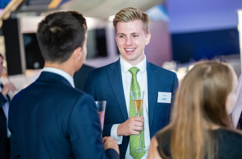 A man in a suit and green tie is smiling while holding a champagne glass at the London conference. He is talking to another man in a suit, with a woman in the foreground, partially visible, perfectly captured by a skilled conference photographer. James Gifford-Mead Photography - Event Photographer London
