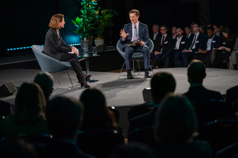 At a London conference, a photographer captures two individuals in formal attire engaged in a panel discussion. The person on the right gestures passionately while speaking, framed by a plant in the background. The attentive audience is partially visible, with some attendees taking notes. James Gifford-Mead Photography - Event Photographer London