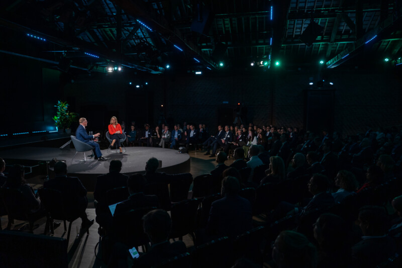 A man and a woman sit on stage during the London Conference, having a discussion in front of a large audience. The venue is dimly lit, with overhead lights casting a blue and green glow. The attendees, potentially captured by a keen conference photographer, are attentively watching the talk. James Gifford-Mead Photography - Event Photographer London