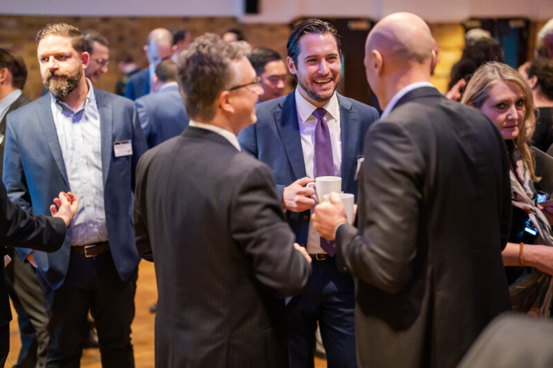 A group of people in business attire are standing and conversing at a London conference. One man in the center holds a white mug, smiling. The background shows more attendees engaged in discussions, capturing the vibrant networking atmosphere through the lens of a skilled photographer. James Gifford-Mead Photography - Event Photographer London