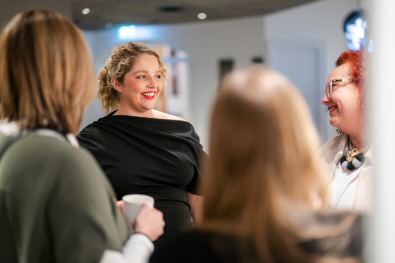 A group of four are engaged in a lively conversation indoors, perhaps at a London conference. One person in a black outfit, possibly the event photographer, is smiling and listening attentively. The setting has the relaxed yet purposeful vibe of a casual gathering or meeting. James Gifford-Mead Photography - Event Photographer London