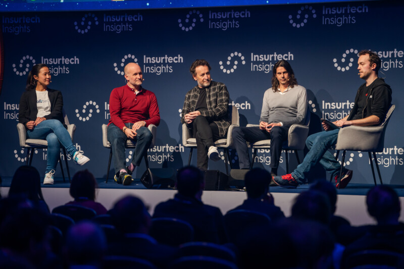 Five people sit in chairs on a stage during a panel discussion at an Insurtech Insights event, expertly captured by a London conference photographer. The backdrop features the event's logo, while an attentive audience fills the foreground, all eyes on the insightful panelists. James Gifford-Mead Photography - Event Photographer London