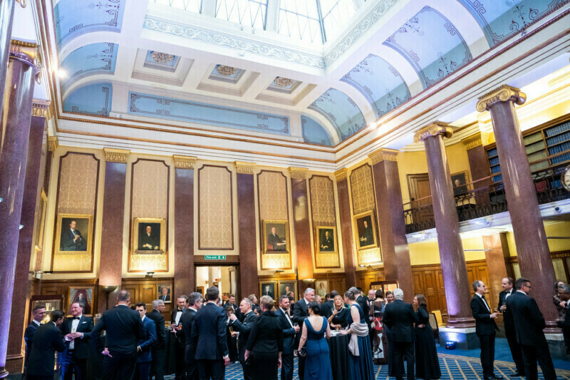 A formal gathering in an elegant hall with high ceilings, ornate decorations, and framed portraits reminiscent of a London conference. Attendees in formal attire mingle and converse under soft lighting as a photographer discreetly captures the moment. A balcony with bookshelves overlooks the scene. James Gifford-Mead Photography - Event Photographer London