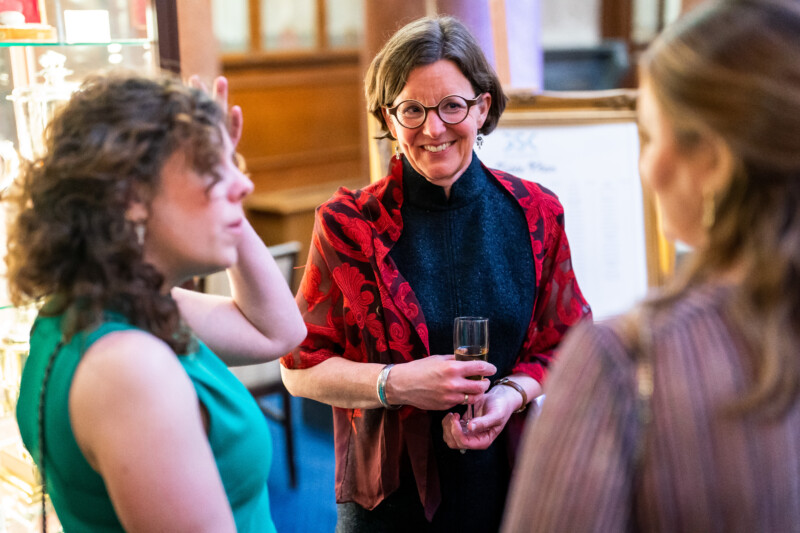 At a London conference, a woman in a red patterned jacket holds a glass and smiles while conversing with two other women. The softly lit background creates a warm and inviting atmosphere, perfectly captured by the talented photographer. James Gifford-Mead Photography - Event Photographer London