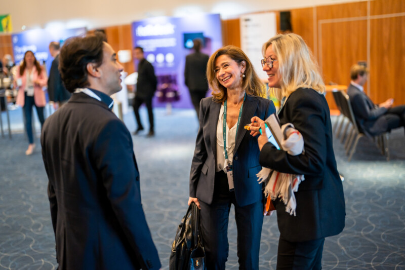Amidst the vibrant scene of a London conference, a photographer captures three professionals engaged in lively conversation. Two women, lanyards and bags in hand, share smiles with a suited gentleman against a bustling backdrop of booths and attendees. James Gifford-Mead Photography - Event Photographer London