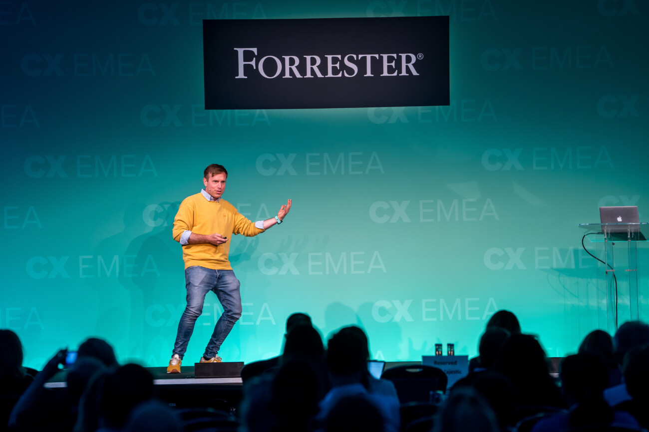 A man in a yellow sweater and blue jeans is speaking energetically on stage at the Forrester CX EMEA conference. Against a teal backdrop, an empty podium stands to the right. Captured by a talented London Conference Photographer, an attentive audience watches from the foreground