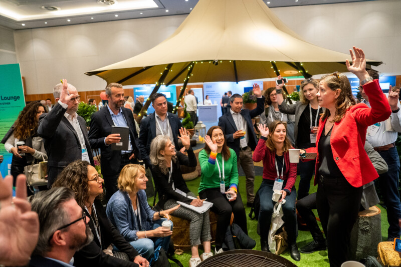 In a conference room beneath a large canopy, a London photographer captures the dynamic scene of people seated and standing, some with raised hands. They are actively engaging in the discussion led by a woman in a red jacket. James Gifford-Mead Photography - Event Photographer London