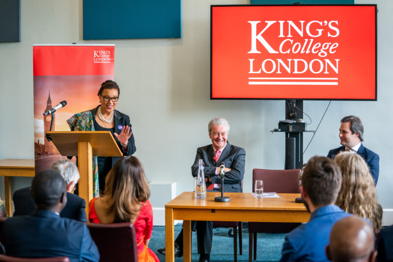 A photographer captures a speaker at a podium addressing an audience at King's College London's conference. A large screen displays the college’s name, while two people are seated at a table on stage, engaging with the attendees. James Gifford-Mead Photography - Event Photographer London