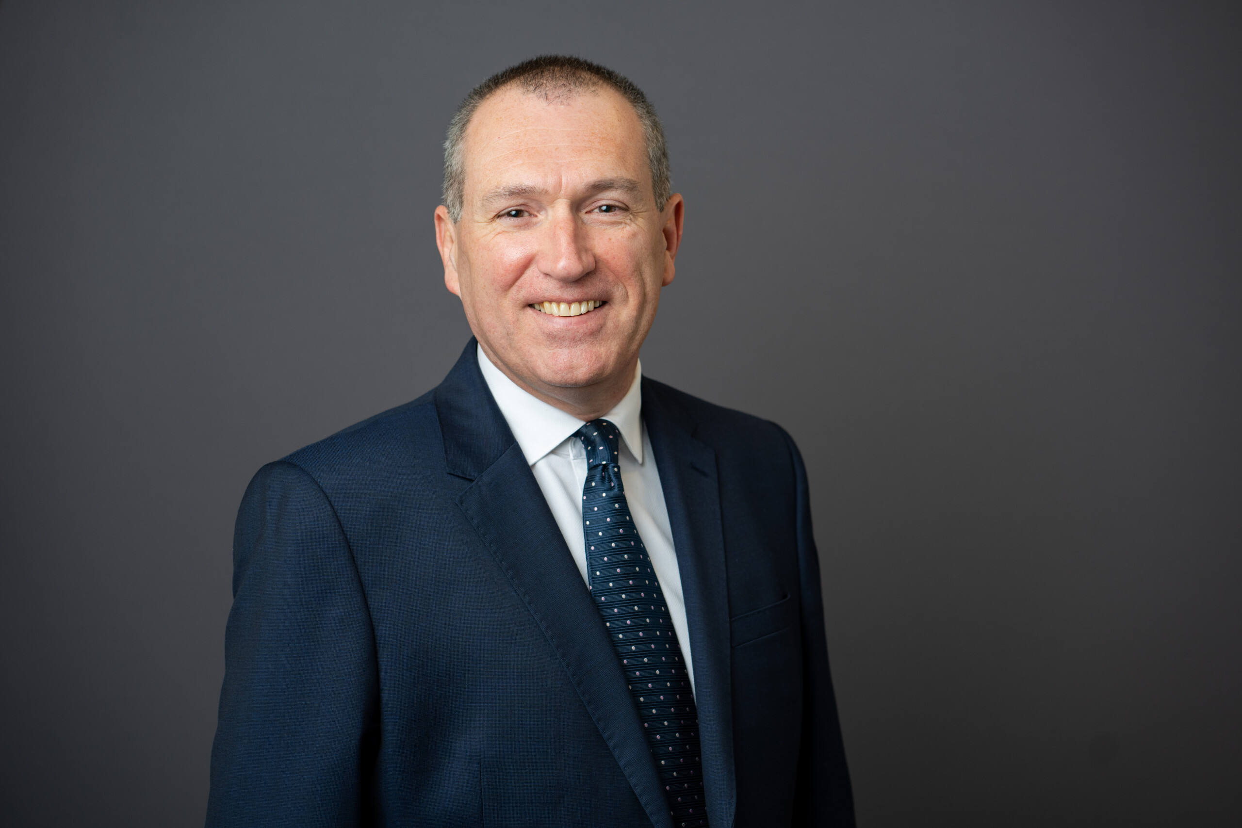 A man in a dark suit and polka dot tie smiles confidently at the camera. His short hair complements the professional aura captured by a skilled corporate headshot photographer, all set against a plain, dark background. James Gifford-Mead Photography - Event Photographer London