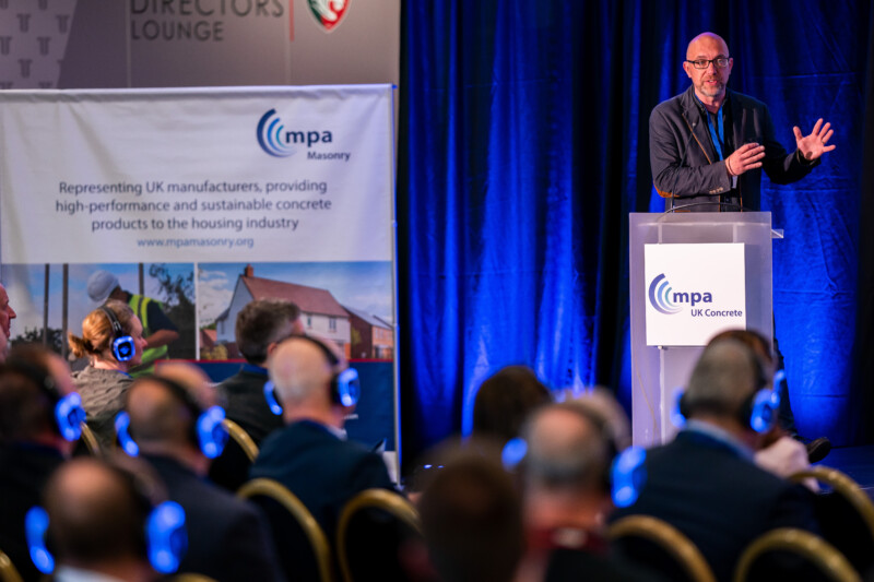 A speaker in a suit and glasses delivers a presentation at a conference while attendees, captured by the London Conference Photographer, wear wireless headphones. A banner behind him promotes UK masonry products for the housing industry as blue lighting illuminates the stage. James Gifford-Mead Photography - Event Photographer London