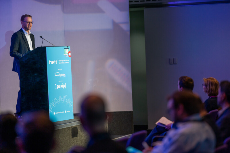 A man in a suit speaks at a podium with a microphone during the London conference. The blue podium, captured by an adept photographer, displays various logos. Several audience members sit facing him, listening attentively as a large screen behind the speaker displays a blurred image. James Gifford-Mead Photography - Event Photographer London